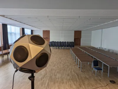 Guľový reproduktor v popredí veľkej sály s akustickým stropom pripravenej na prednášky