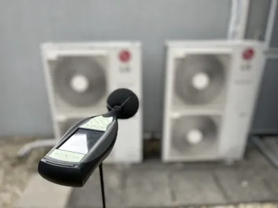 Zvukomer v popredí meria hluk dvoch vonkajších klimatizačných jednotiek na fasáde budovy.