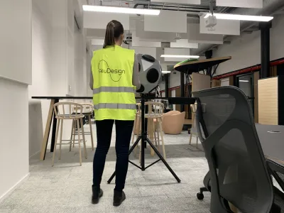 Pracovníčka AkuDesign nastavuje meracie zariadenie na statíve v modernej kancelárii s vysokými stolmi.