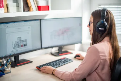 Žena so slúchadlami analyzuje pôdorys budovy na dvoch monitoroch pri akustickom posudzovaní.