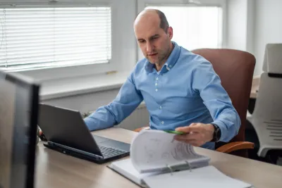 A man at a laptop checks printed technical documentation in an office.