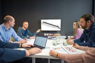 Tím pri stole diskutuje o technickom výkrese oceľovej konštrukcie so schodiskom na obrazovke.